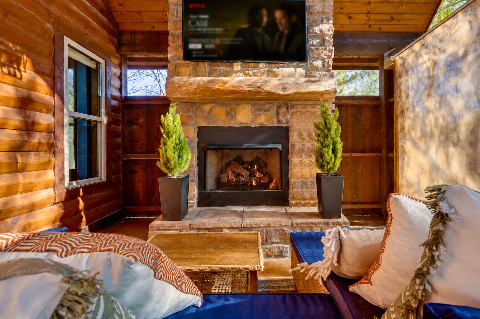 A log cabin interior with a stone fireplace, a television mounted on the wall, and a cozy seating area with cushions.