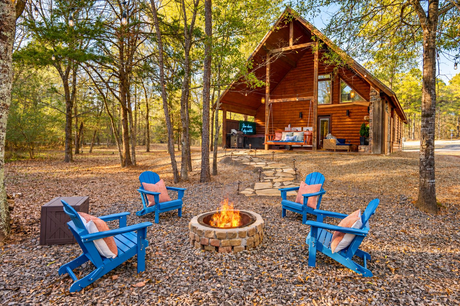 A cabin with a stone fire pit surrounded by blue Adirondack chairs, set in a forested area.