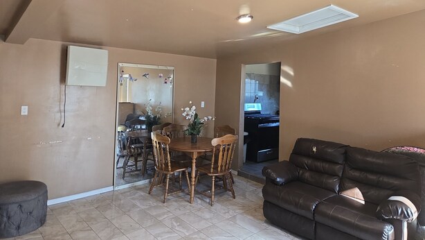 A living room with a brown leather sofa, a dining table with chairs, and a kitchen visible through an open doorway.