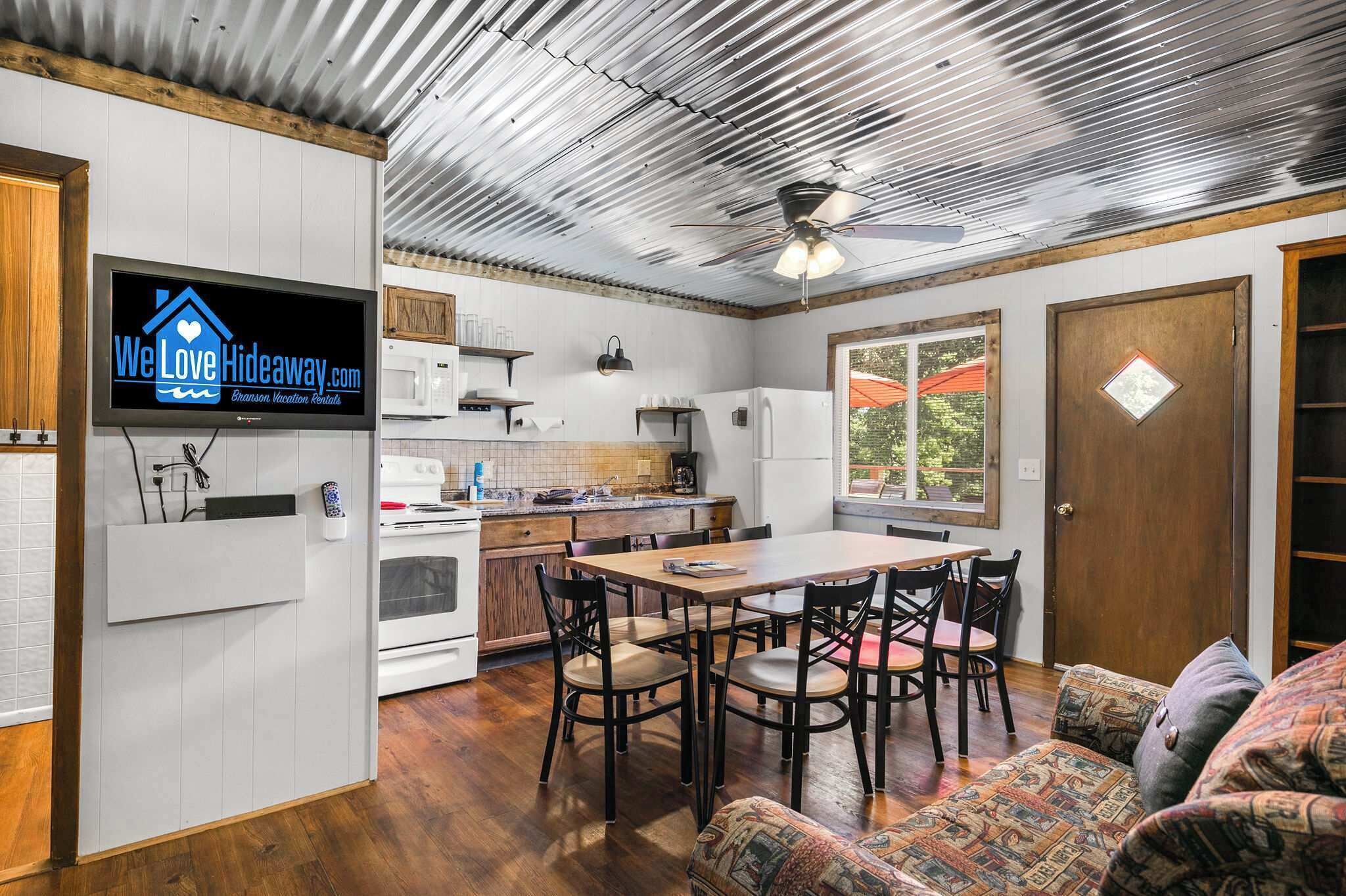 A kitchen with a dining area, a television displaying 'We Love Hideaway.com,' and a ceiling fan.