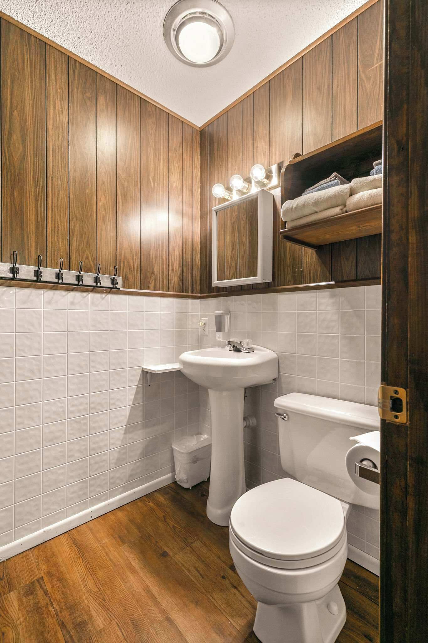 A bathroom with wooden paneling, a white sink, a toilet, and a vanity mirror with lights.
