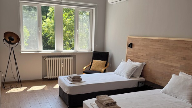 Una camera da letto con due letti, una finestra vista alberi, una lampada da terra e una ventola a soffitto.