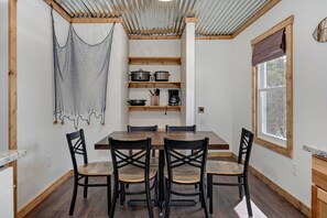 A dining area with a wooden table and chairs, a hanging net, and shelves with kitchen items. - Blue Heron · Lakefront Cabin · Pool & Dock (Galena)