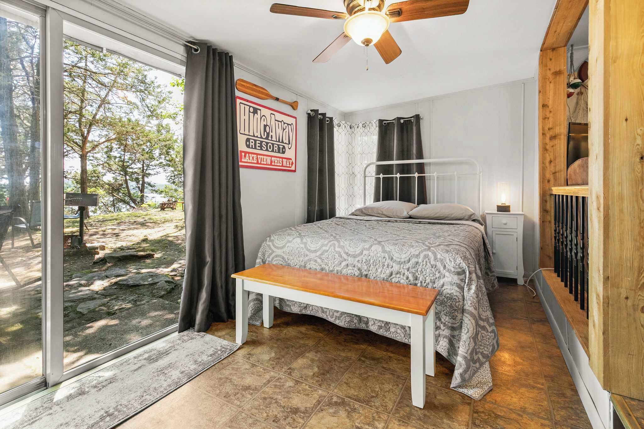 A bedroom with a bed, a bench, a nightstand, a ceiling fan, and a view of the outdoors.