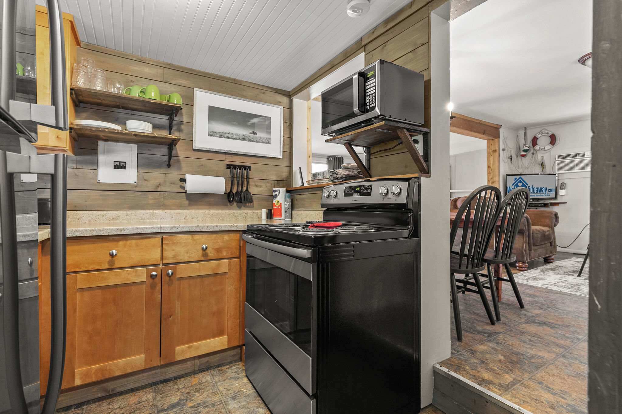 A compact kitchen with a stove, microwave, wooden cabinets, and a dining area with a chair.