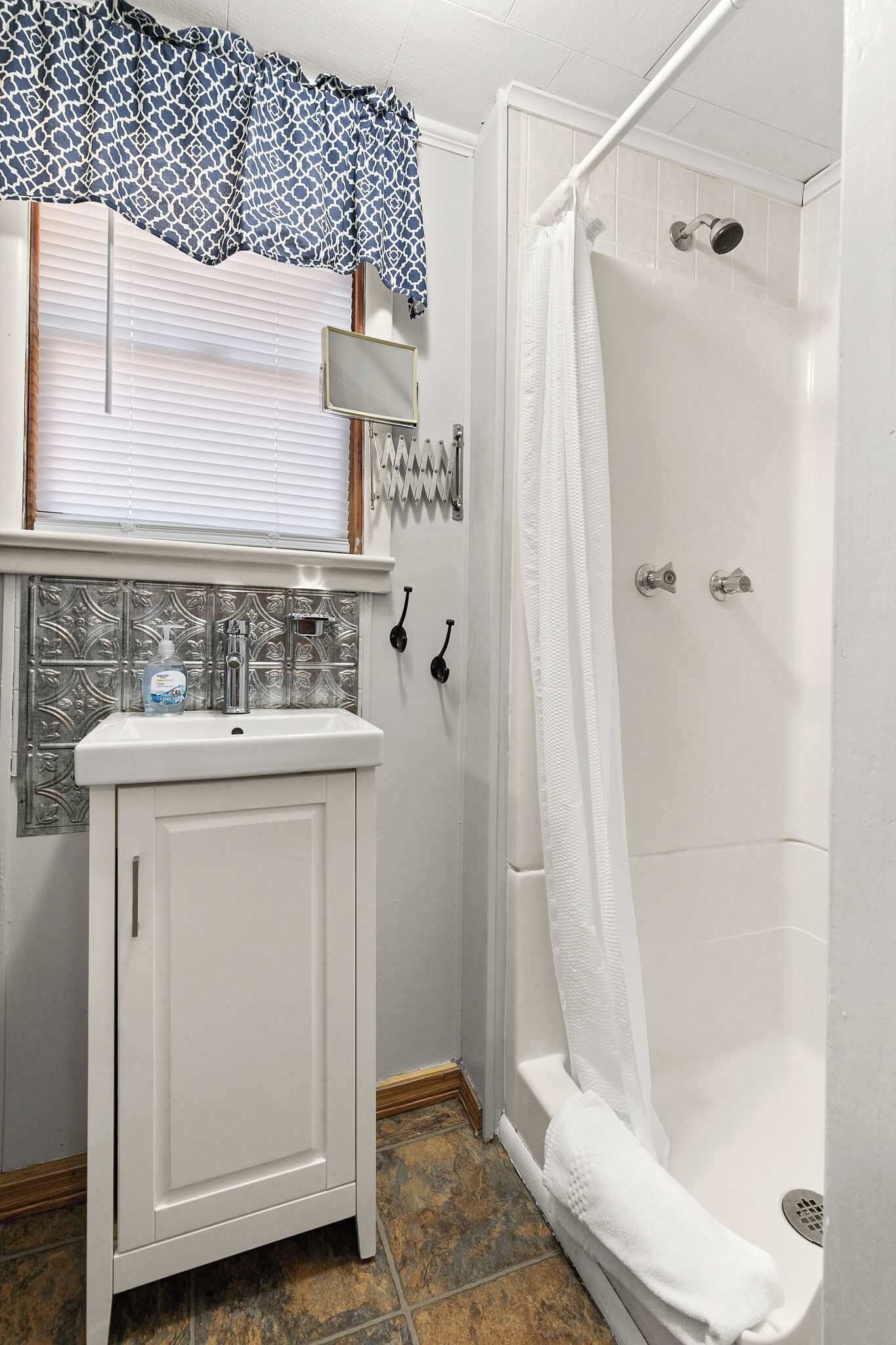 A bathroom with a shower, a sink with a soap dispenser, and a window with blinds.