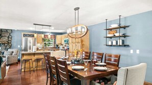 Un coin repas avec une table en bois dressée pour quatre personnes, entourée de chaises, une cuisine en arrière-plan et un mur en pierre.