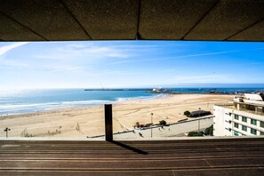 Blick auf einen Strand vom Gebäude aus, mit einer Holzterrasse, Sand, dem Meer und einem entfernten Schiff.