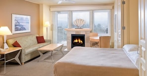 Una habitación de hotel con cama, sofá, mesa, chimenea y vistas a la ciudad a través de amplios ventanales.