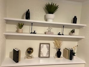 Una estantería blanca con adornos como libros, un cuadro enmarcado y plantas en macetas.
