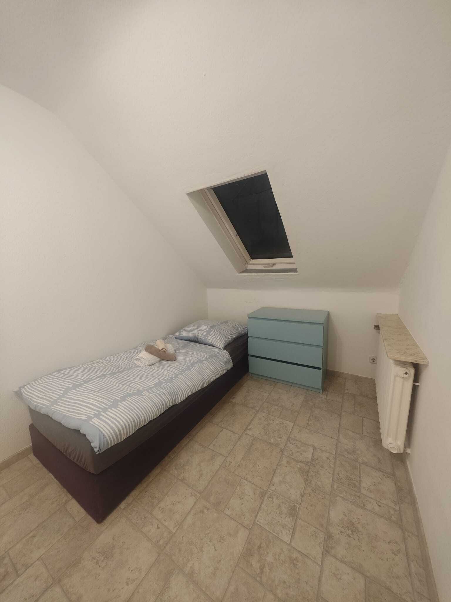 Una habitación pequeña con cama individual, techo inclinado, ventana, mesita de noche y radiador.