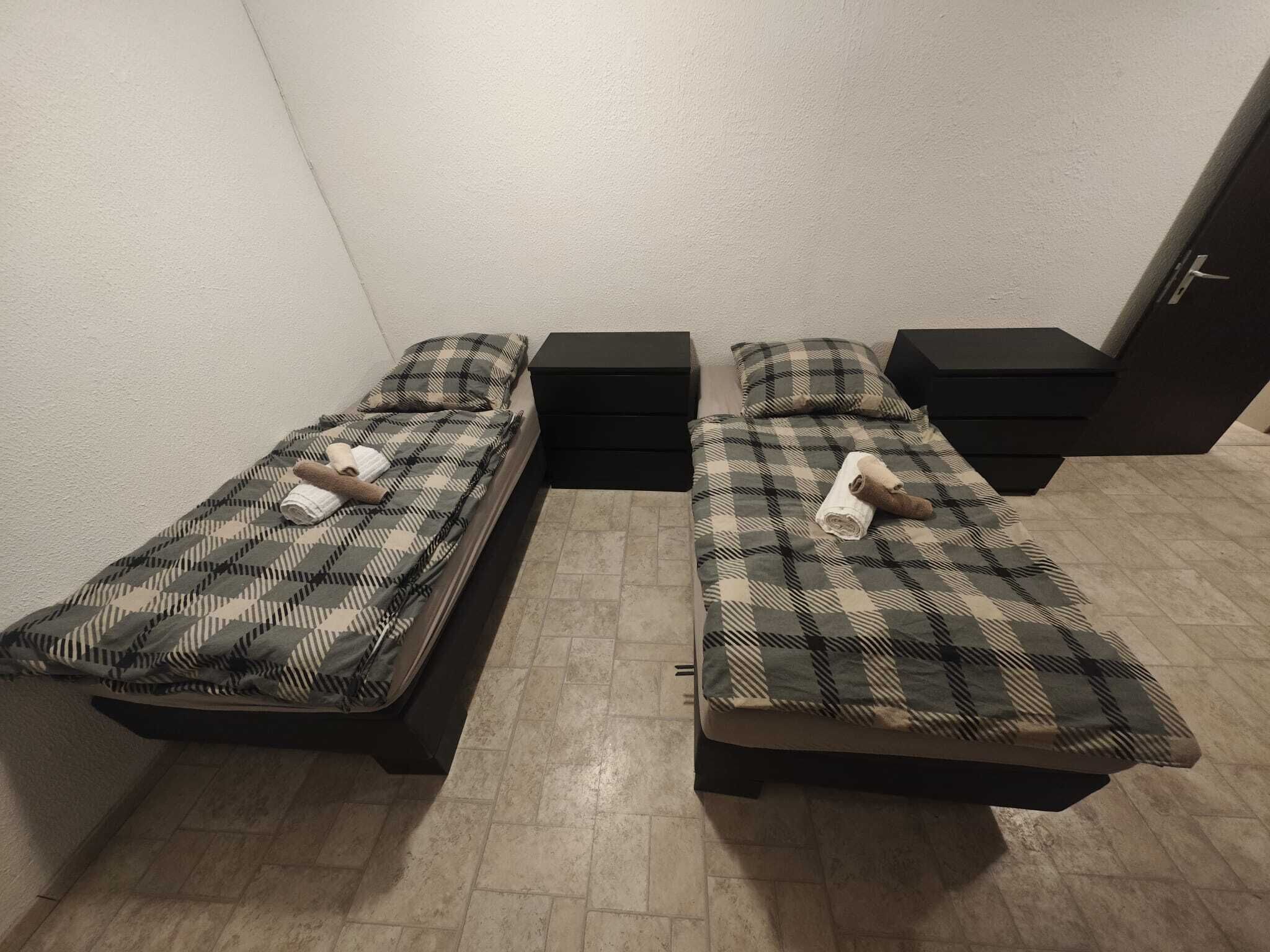 Twee eenpersoonsbedden met geruite beddengoed, zwarte nachtkastjes en een betegelde vloer in een kleine kamer.