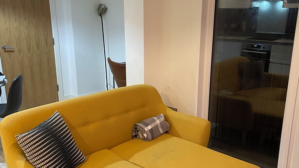 En moderne stue med en gul sectional sofa, en lille skammel og en glasdør til et køkken.