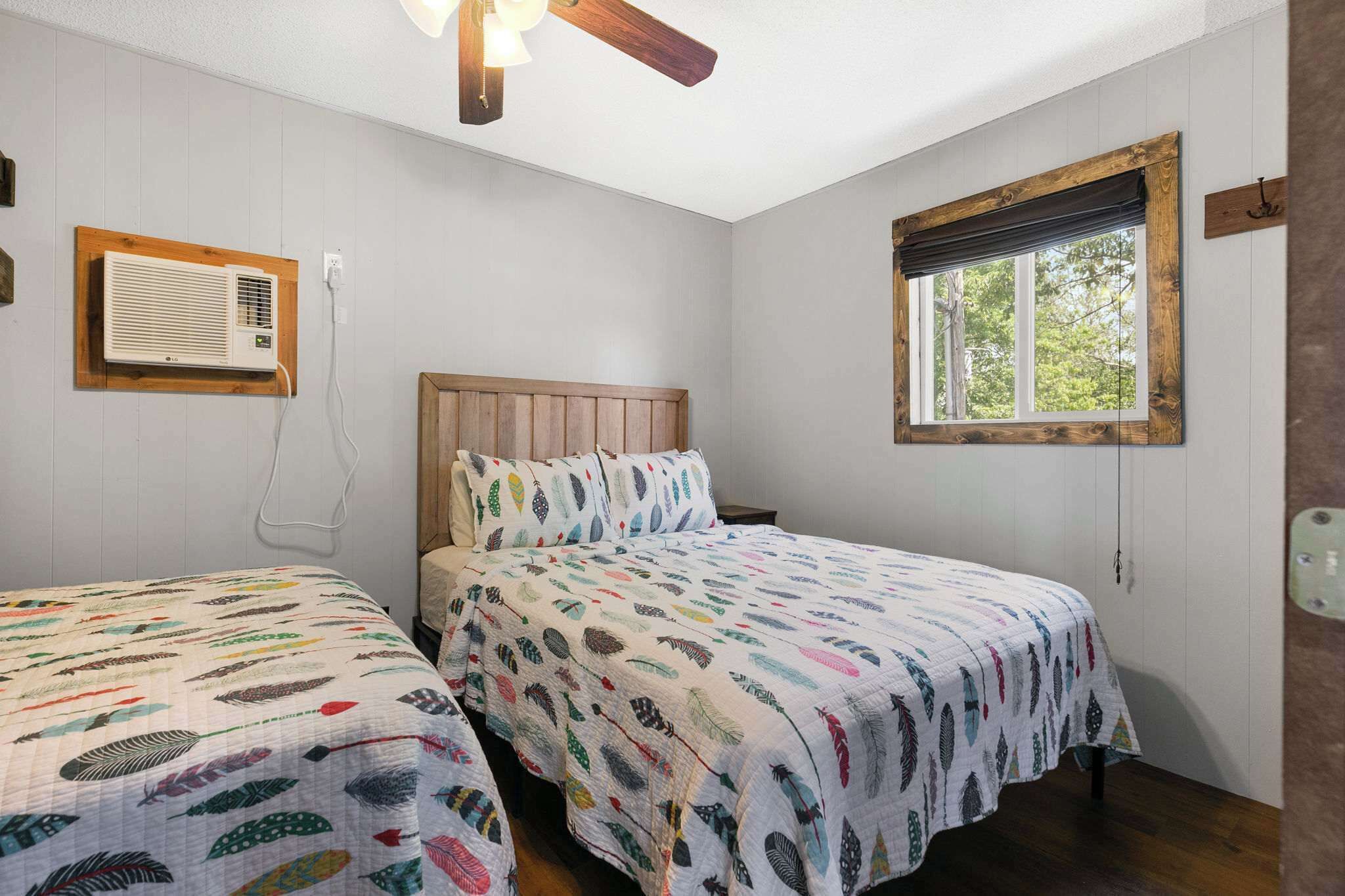 A bedroom with two beds, a ceiling fan, an air conditioning unit, a window with a view, and a wooden headboard.