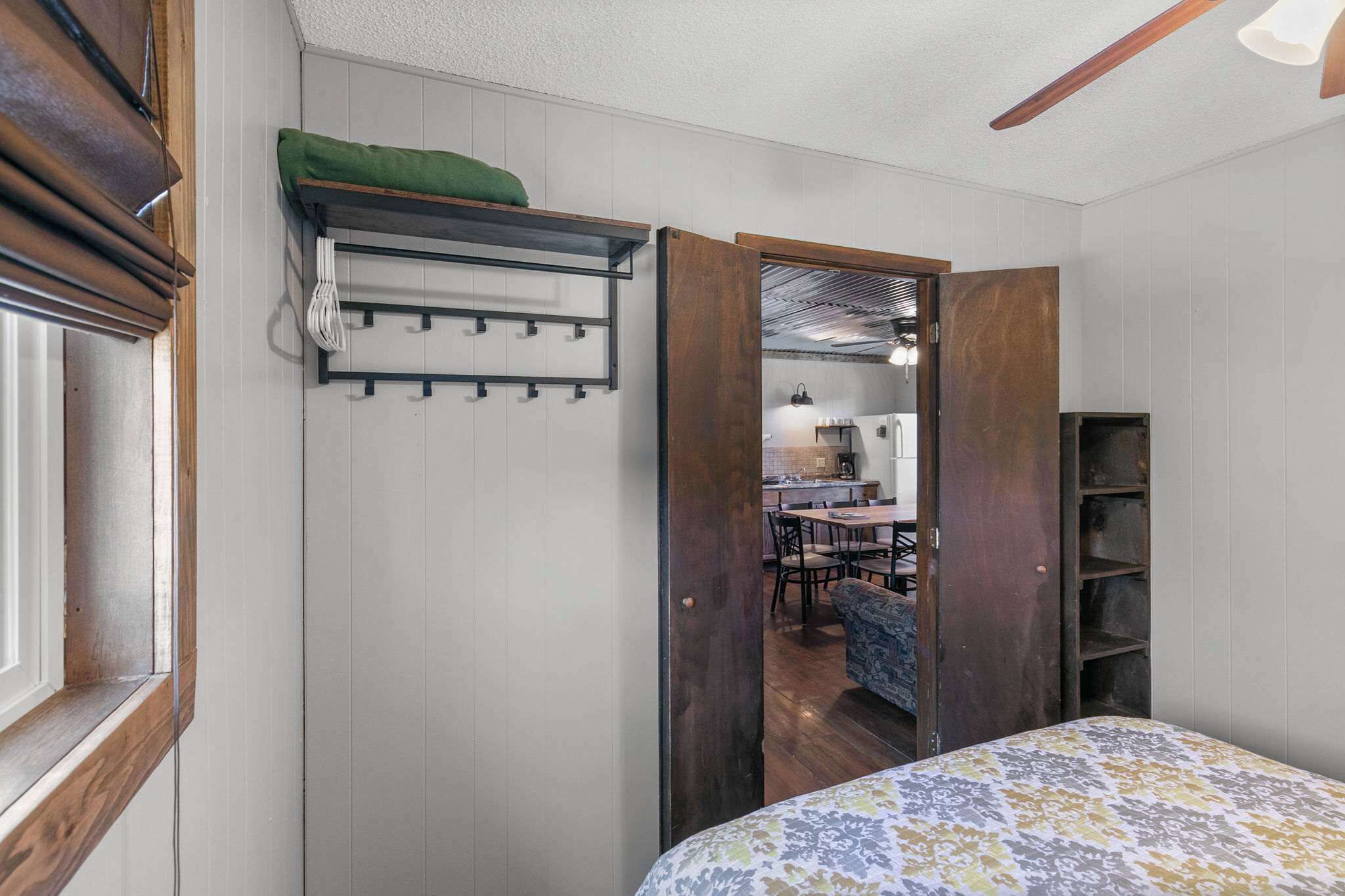 Une pièce comprenant un lit, une porte en bois donnant sur une cuisine, un porte-manteau et une petite étagère.