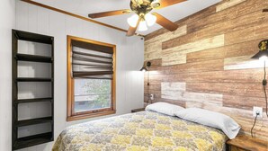 Une chambre avec un mur en bois, un ventilateur de plafond, un lit recouvert d’une courtepointe à motifs, une fenêtre avec des stores et une étagère.