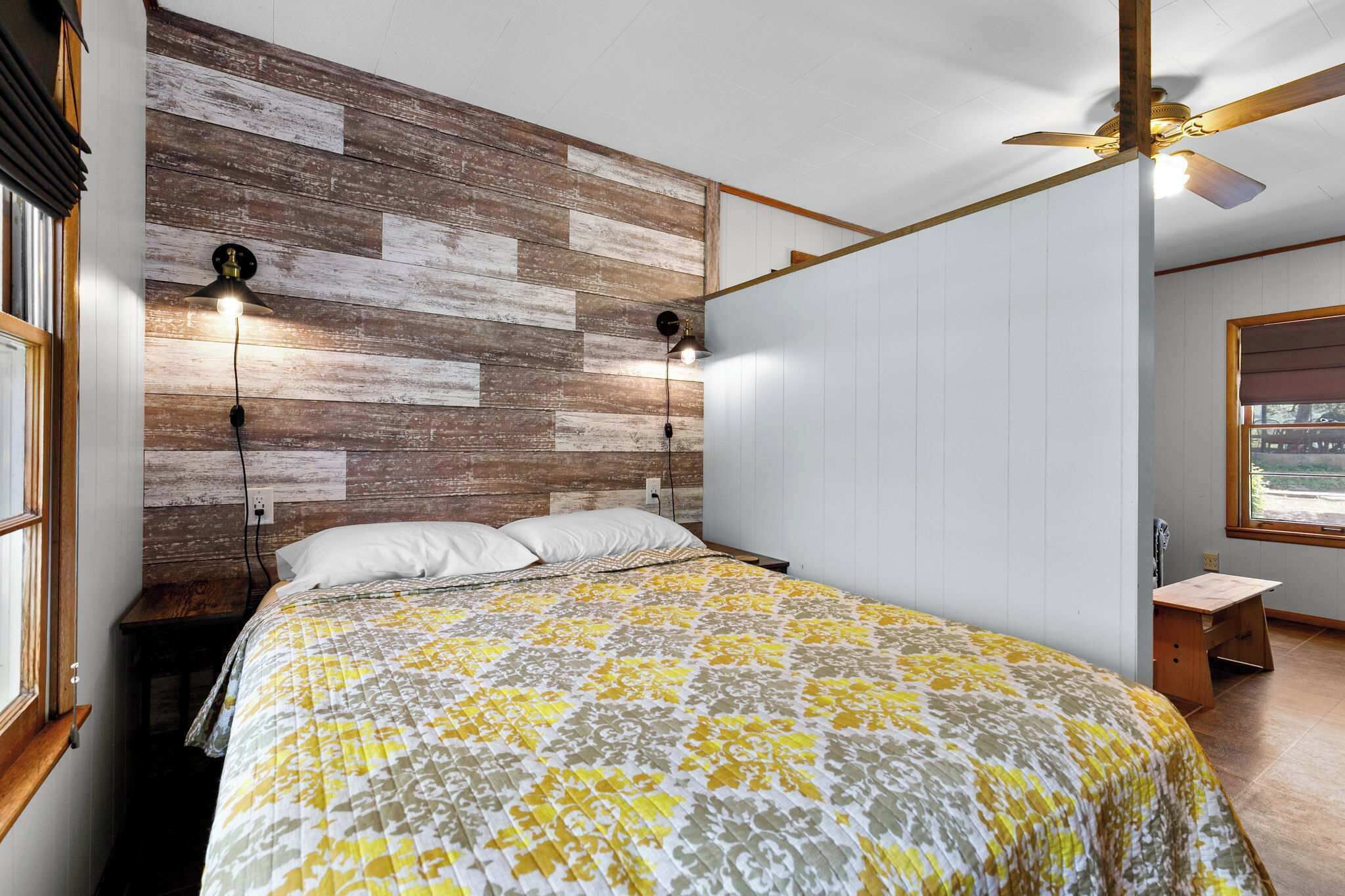 Een slaapkamer met een houten hoofdbord, een bed met een geborduurde spreideken, twee wandlampen, een plafondventilator en een raam met uitzicht.
