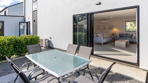 Un pati exterior modern amb taula i cadires de vidre, una porta corredissa que dona a un espai interior i un arbust al fons.