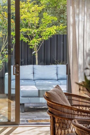 Ein moderner Außenbereich mit Sitzgelegenheiten, darunter eine Couch und Korbstühle, eine Glastür, die zu einem Garten führt, und ein schwarzer Zaun im Hintergrund.