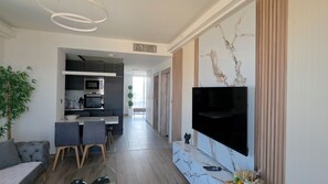 Un salon moderne doté d’un téléviseur à écran plat, d’un coin repas avec une table et des chaises, et d’une cuisine équipée d’appareils électroménagers intégrés.