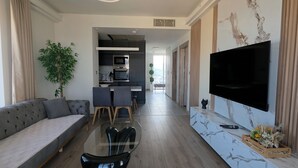 Ein modernes Wohnzimmer mit einer Couch, einem Esstisch, Stühlen, einem Fernseher und einer Küche im Hintergrund.