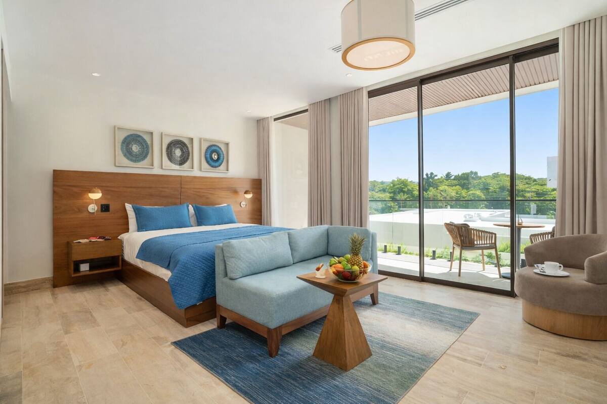 A modern hotel room with a large bed, a seating area with a fruit bowl, a balcony with a view, and a large window.