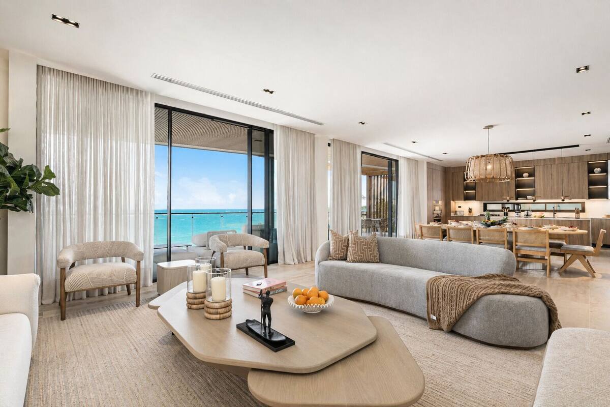 A modern living room with a large window overlooking the ocean, a grey sofa, a coffee table with decorative items, and a dining area with wooden furniture.