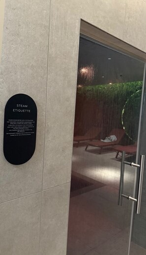 A steam room with a sign detailing etiquette, a glass door, and a view of a lounge area with chairs and a green wall. - Bespoke Piccadilly 2-Bed Retreat Gym Spa Pool (Manchester)