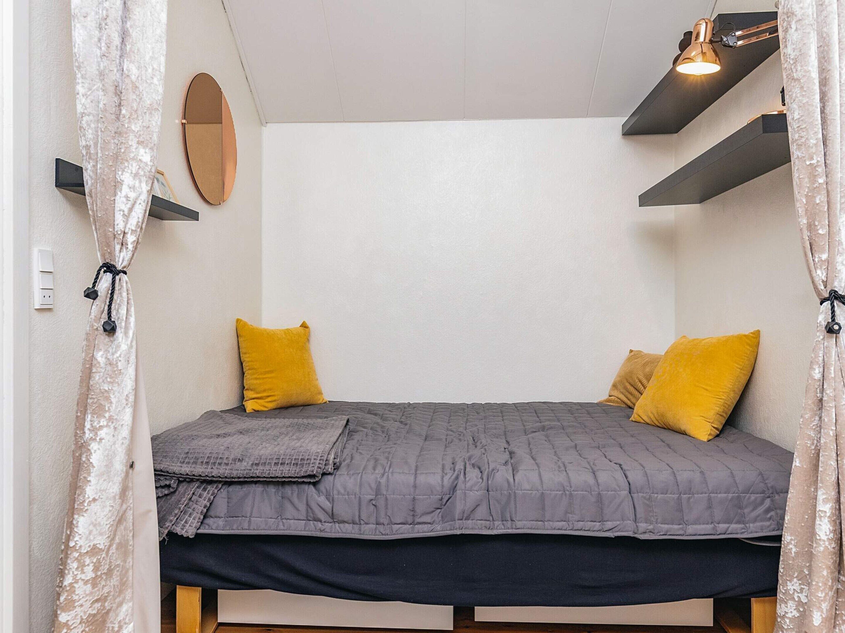 A small, cozy bedroom with a bed, two pillows, a bedside lamp, a mirror, and a shelf above the bed.