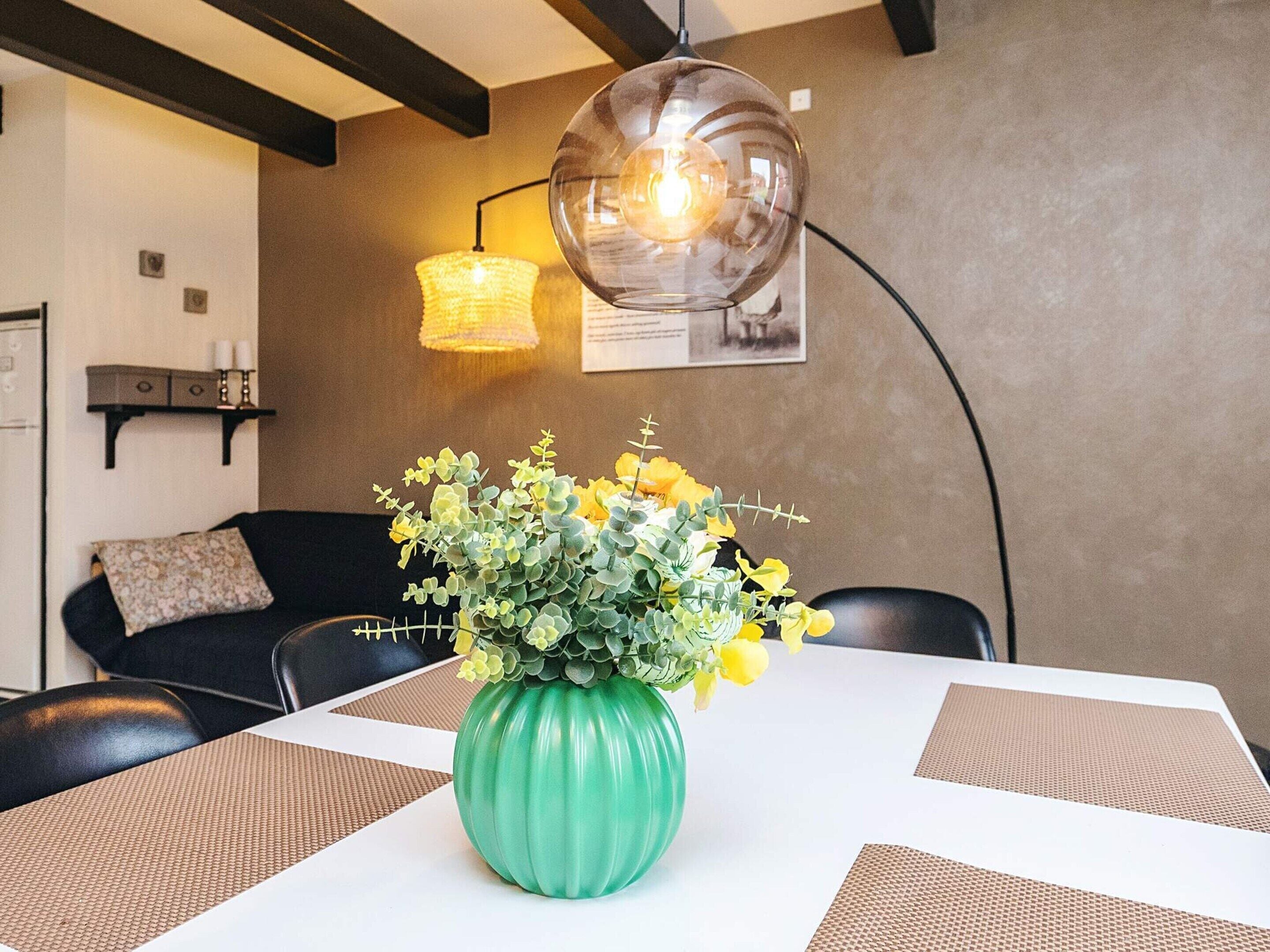 Een eethoek met een tafel, stoelen, een groene vaas met gele bloemen, een hanglamp en een wandplank met kaarsen.