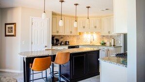 A kitchen with a central island, bar stools, pendant lighting, and a backsplash. - Rexford Sound HideAway-Pool and Pet Friendly (Navarre)