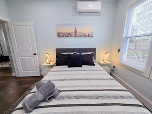 Ett sovrum med en säng, två sängbord med lampor, en målning som föreställer en stadsbild och en luftkonditioneringsanläggning.