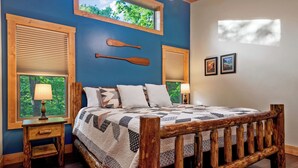 Quarto com estrutura de cama de madeira, duas janelas com persianas, colcha acolchoada e duas luminárias fixadas na parede.