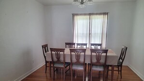 Uma sala de jantar com uma mesa retangular e seis cadeiras, um ventilador de teto e uma janela com cortinas.