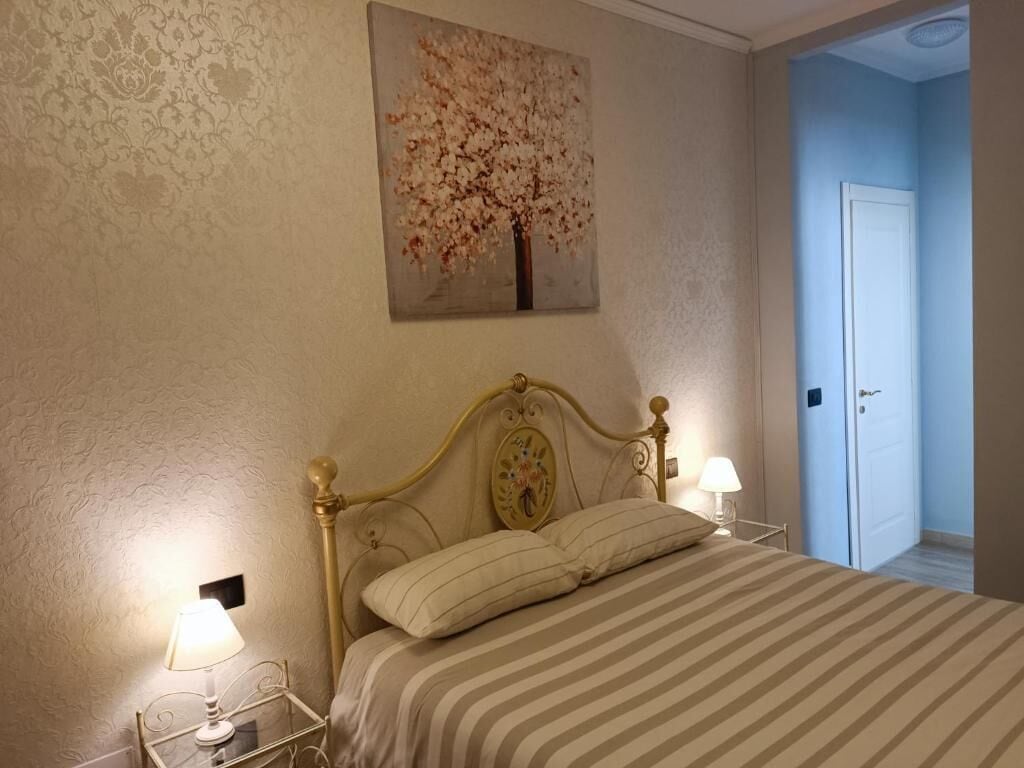 A bedroom with a bed, two bedside lamps, a wall-mounted painting of a tree, and a door to another room.