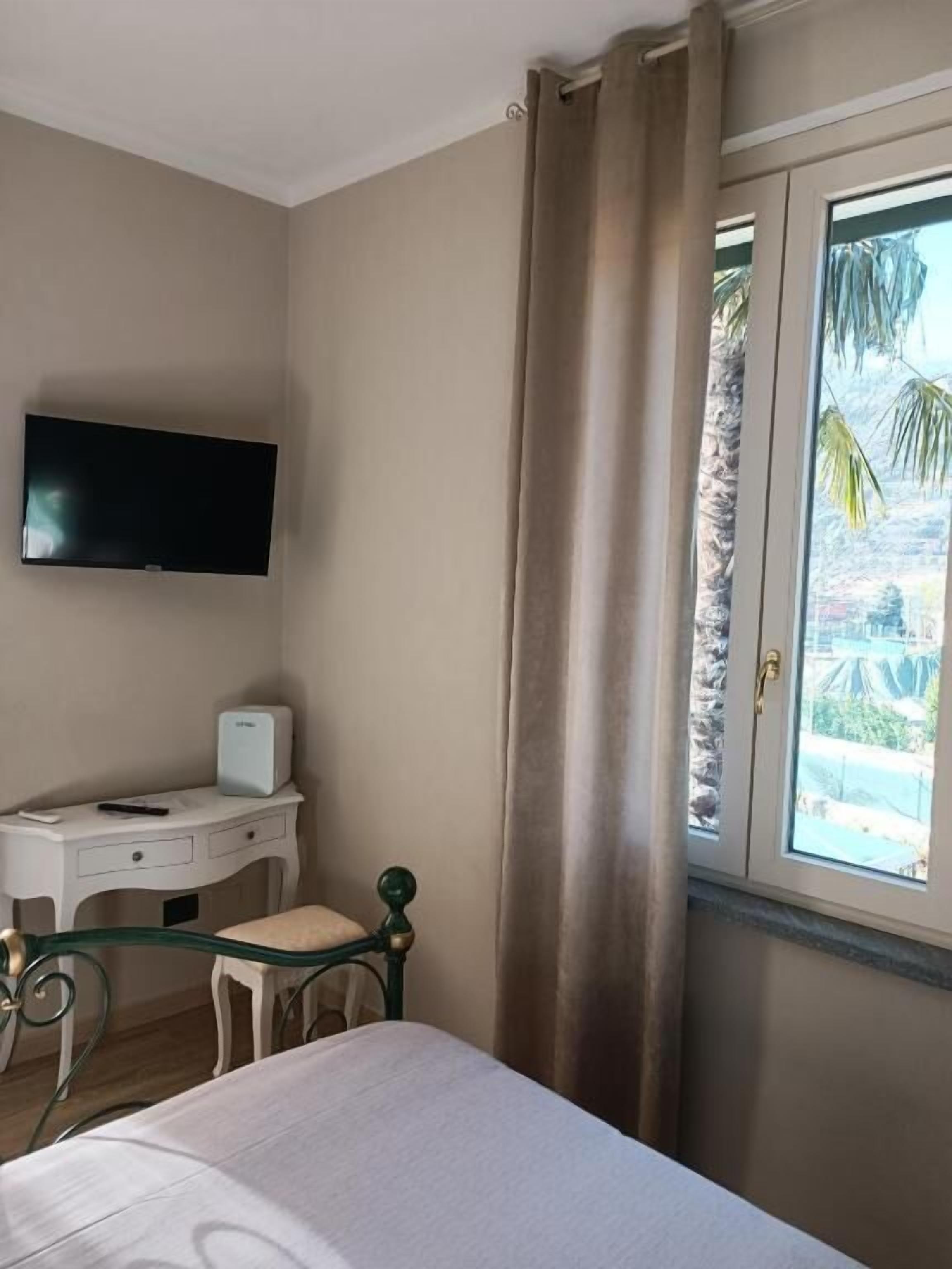 Ein Schlafzimmer mit Bett, Schreibtisch mit Fernseher, Fenster mit Vorhängen und Blick auf Palmen.