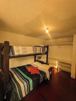 Habitación con literas, cabecera de madera, mesita de noche con lámpara y estante con ropa colgada.