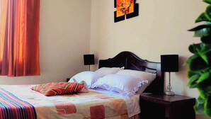 A bed with a colorful blanket, two bedside lamps, a wall art piece, and a window with curtains. - Sumaq Wasi Inca Trails (Lima)