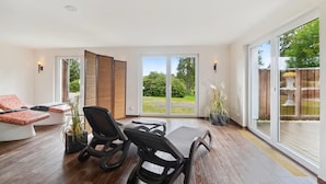 A spacious room with a large sliding glass door leading to a garden, a dining table with chairs, and a sofa with cushions. - Blockhaus im Harz fur Familien (Oberharz am Brocken)