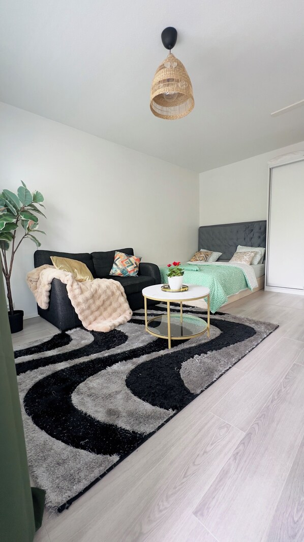 A modern bedroom with a black and white rug, a round coffee table, a sofa with cushions, a bed with a green bedspread, and a hanging light fixture.