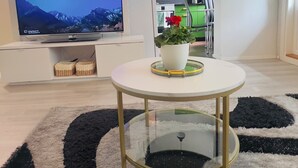 A modern living room with a flat-screen TV, a round glass coffee table with a white base, a black and white rug, and a kitchen in the background. - Modern Apartment in Myyrmäki – Direct Train to Helsinki & Airport (Vantaa)