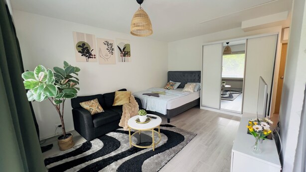 A modern bedroom with a bed, a black sofa, a small round table, a potted plant, and artwork on the wall. - Modern Apartment in Myyrmäki – Direct Train to Helsinki & Airport (Vantaa)