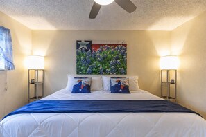 A hotel room with a bed, two bedside lamps, a ceiling fan, and a wall painting.