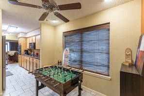 A room with a foosball table, a ceiling fan, a kitchen area, and a window with blinds. - Waffle House | Pool · Game Room · Putting Green · (Abilene)