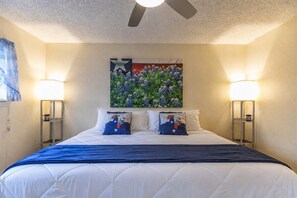 A hotel room with a bed, two bedside lamps, a ceiling fan, and a wall-mounted artwork.