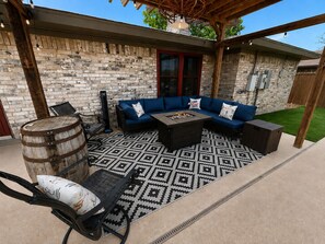A covered patio with a stone wall, a blue sofa set, a fire pit, and a metal chair. - Waffle House | Pool · Game Room · Putting Green · Walk to ACU  · 🤠 (Abilene)