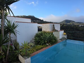 A pool with a stone wall and plants, a white building with a red roof, and a view of hills in the background. - Ocean Heated Pool Jacuzzi View I Flat (Santa Cruz)