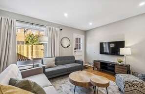 A modern living room with a grey sofa, a round coffee table, a TV, and a window with curtains. - Staycation in Del Rey 10 min to DC Firepit Crib (Alexandria)