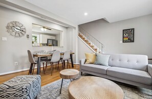 A modern living room with a grey sofa, a dining table with chairs, a round coffee table, and a staircase. - Staycation in Del Rey 10 min to DC Firepit Crib (Alexandria)
