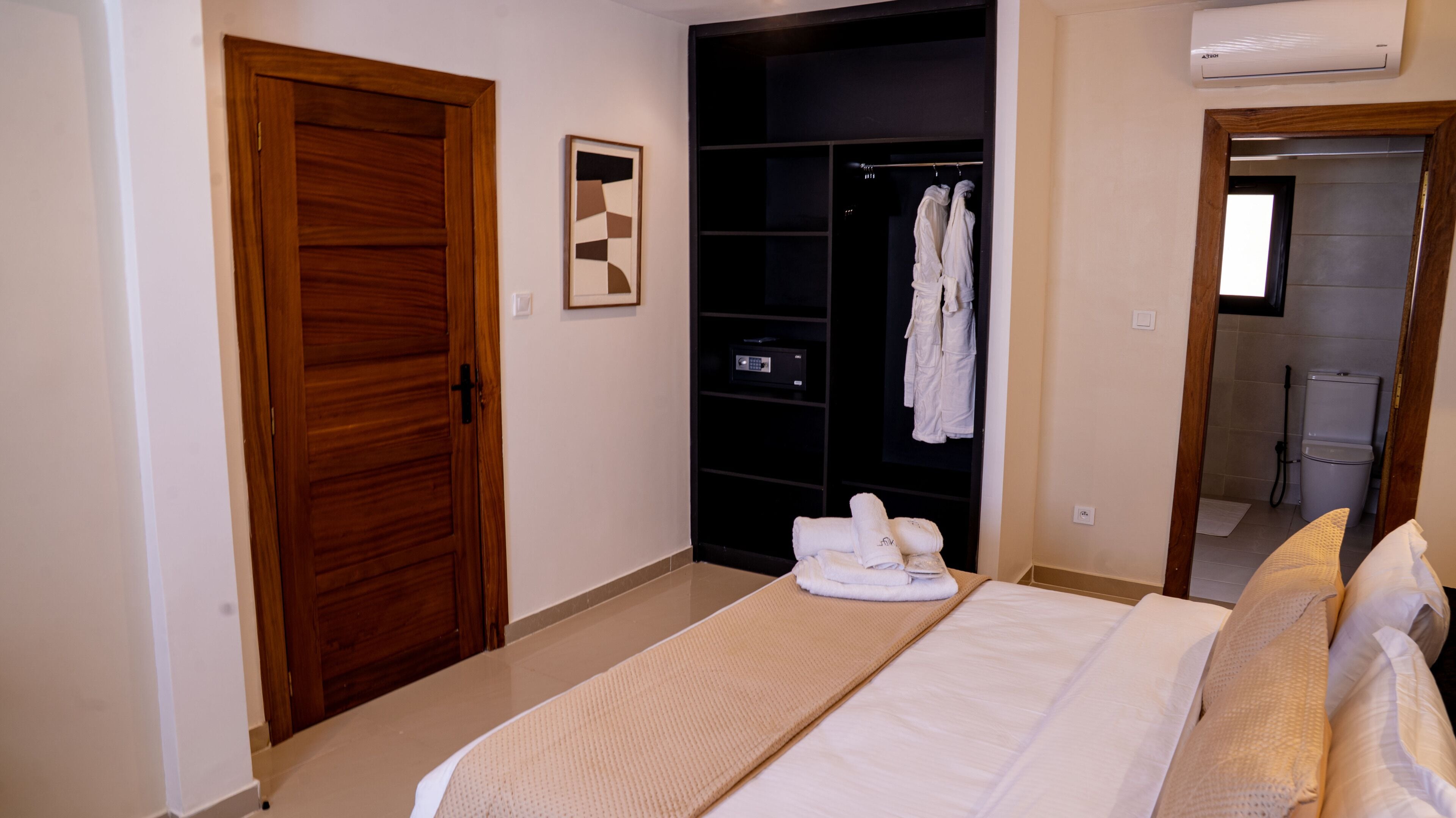 A bedroom with a wooden door, a closet with hanging robes, a bed with white linens, and a bathroom visible through an open door.